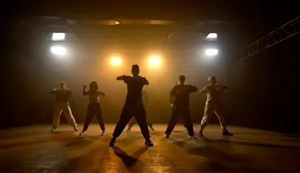A group of energetic hip-hop dancers in a variety of styles dance on an expansive stage illuminated by bright neon lights, their silhouettes surrounded by a halo of sidelights. The wide-format cinematography captures their neat steps, vibrant energy and youthful expressions. The fast-paced cinematography echoes the beat of the music, demonstrating the complexity of the footwork, the explosiveness, and the teamwork.