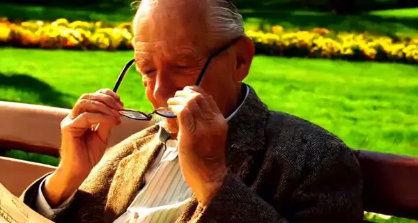 An old man adjusts his glasses while reading a newspaper in the sunlit park